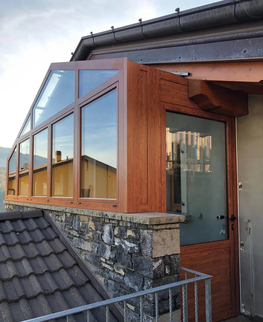 Veranda con vetrate e porta in profilo effetto legno su muretto in pietra, accanto a tetto in tegole grigie e ringhiera